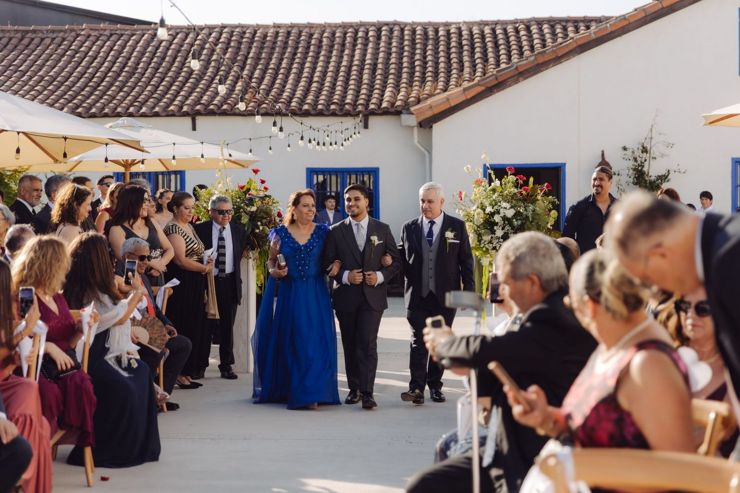 Groom escorted by parents down outdoor wedding aisle lined with seated guests.