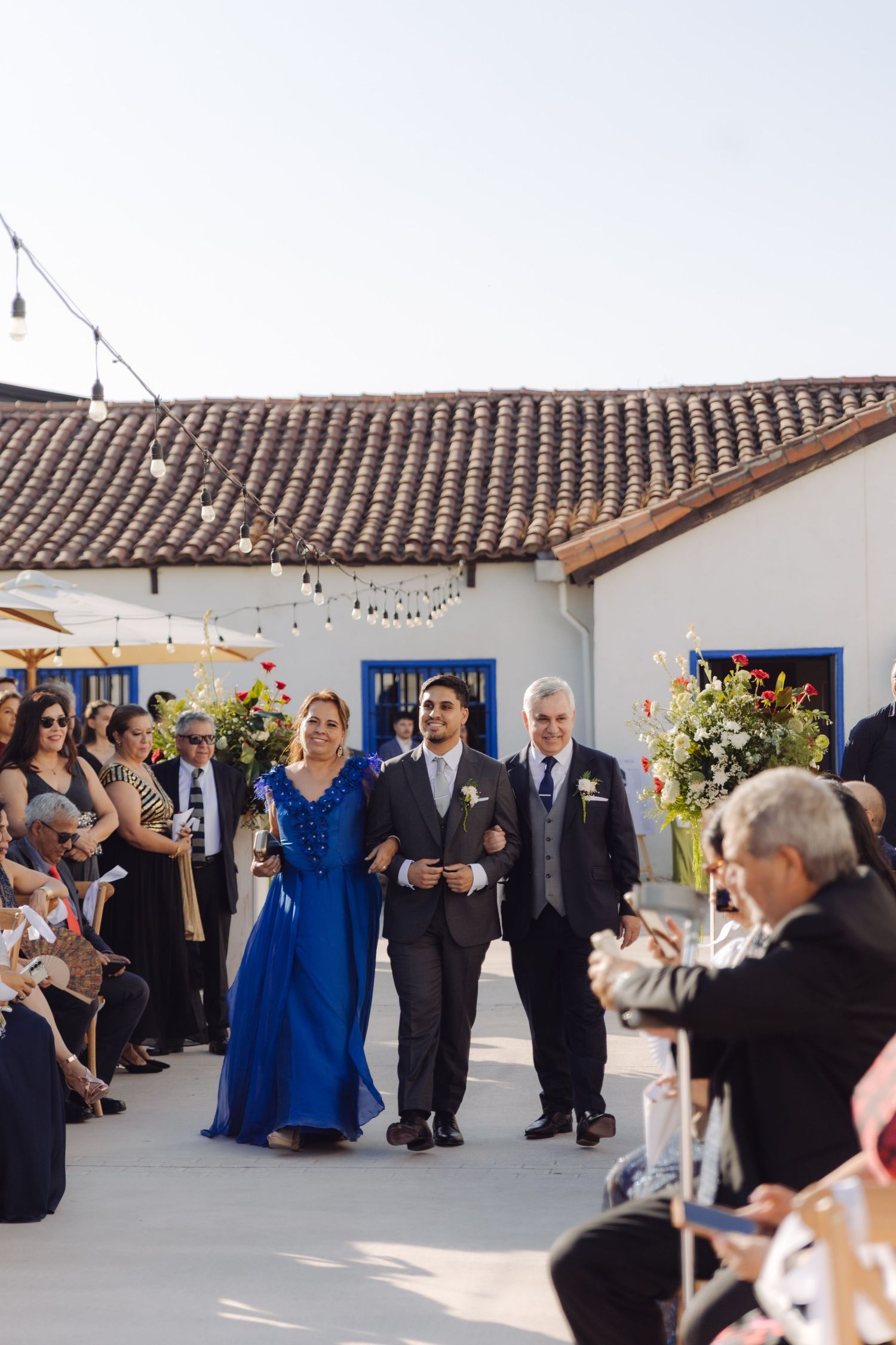 Groom walks down outdoor wedding aisle with parents in blue and navy attire.
