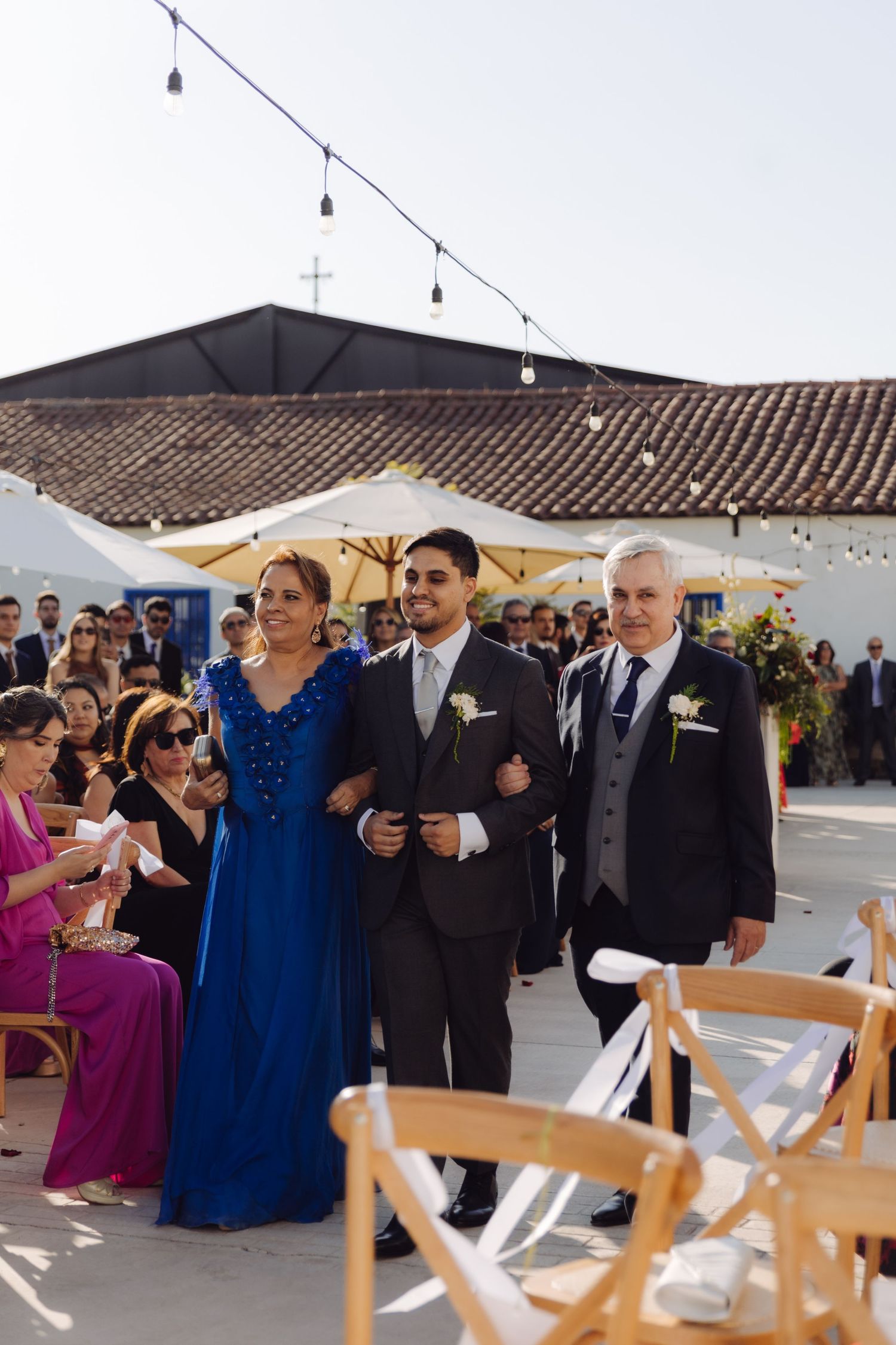 Groom in gray suit walks aisle with mother in royal blue dress and father.