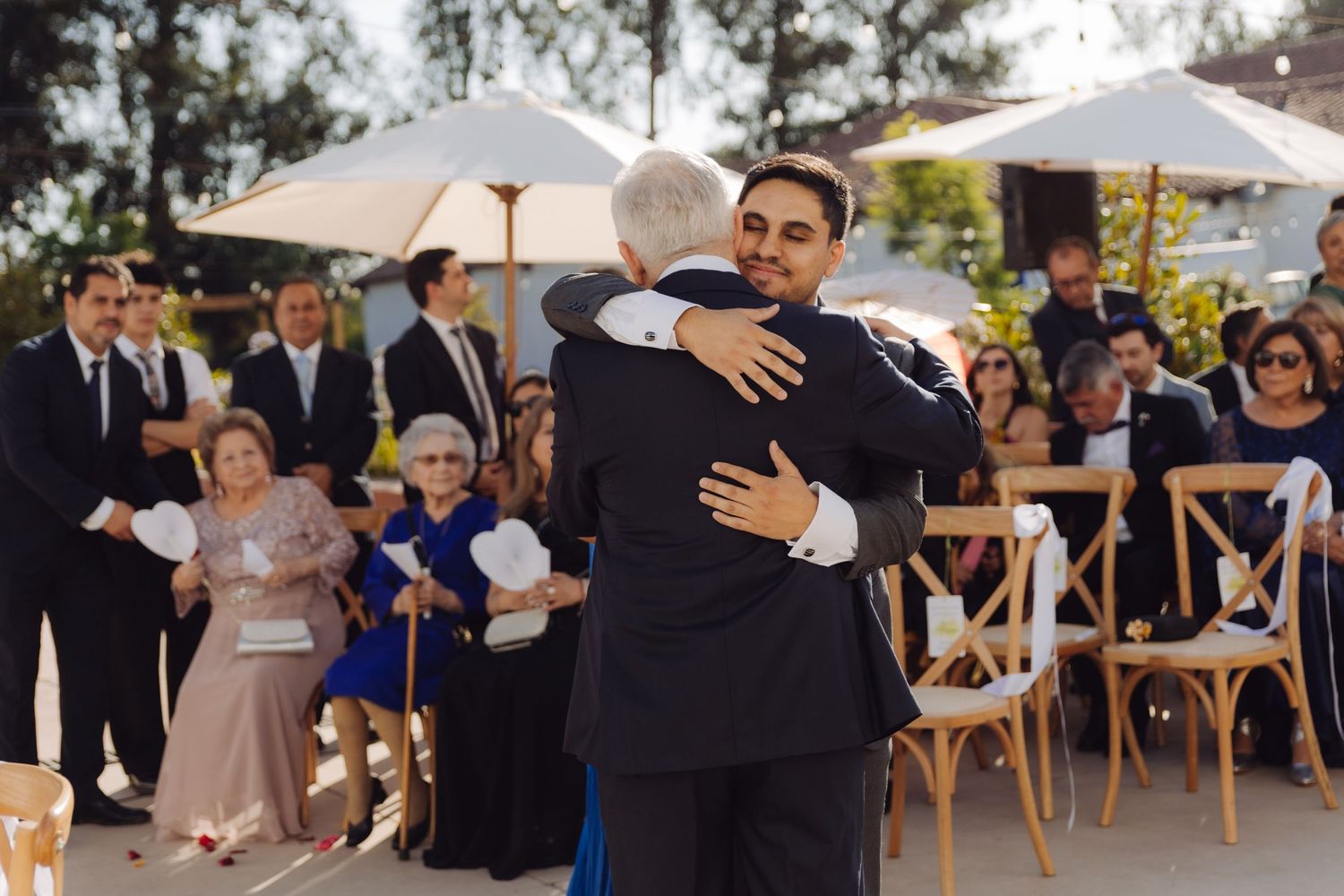 Groom and father share emotional hug during outdoor wedding ceremony.