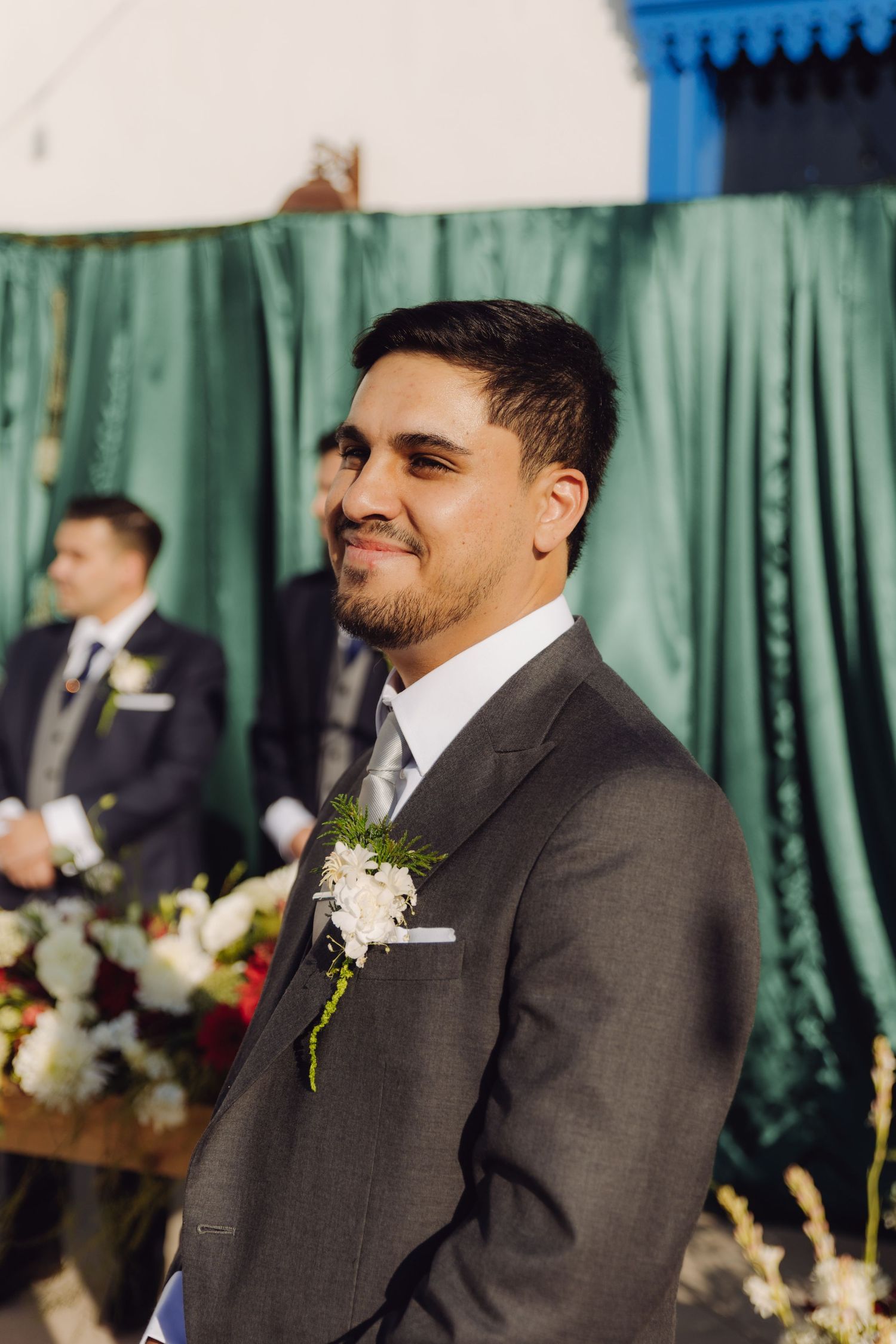 Groom in gray suit with boutonniere smiling at altar during wedding ceremony.