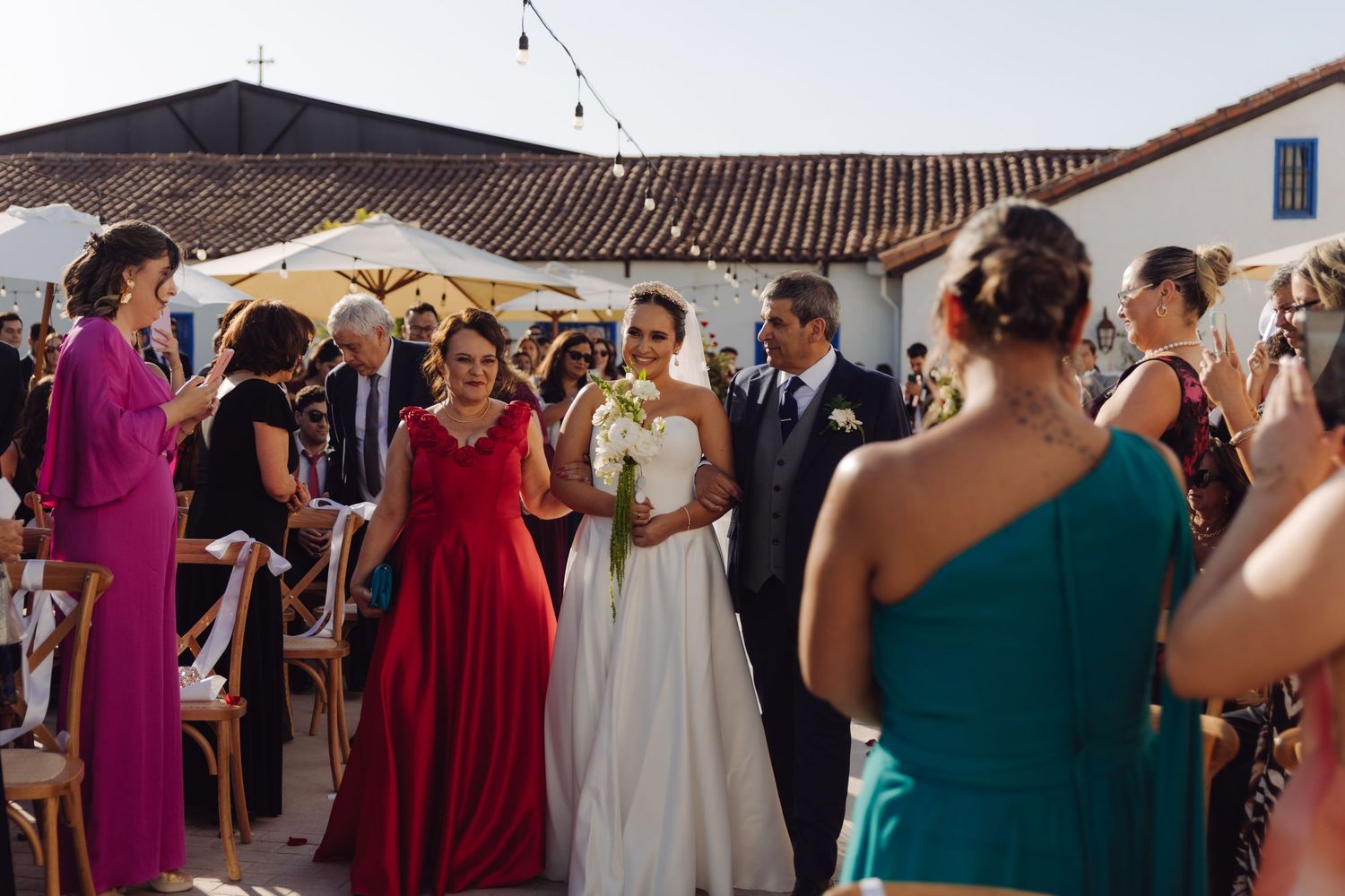Bride in white dress walks down outdoor aisle with parents at wedding ceremony.