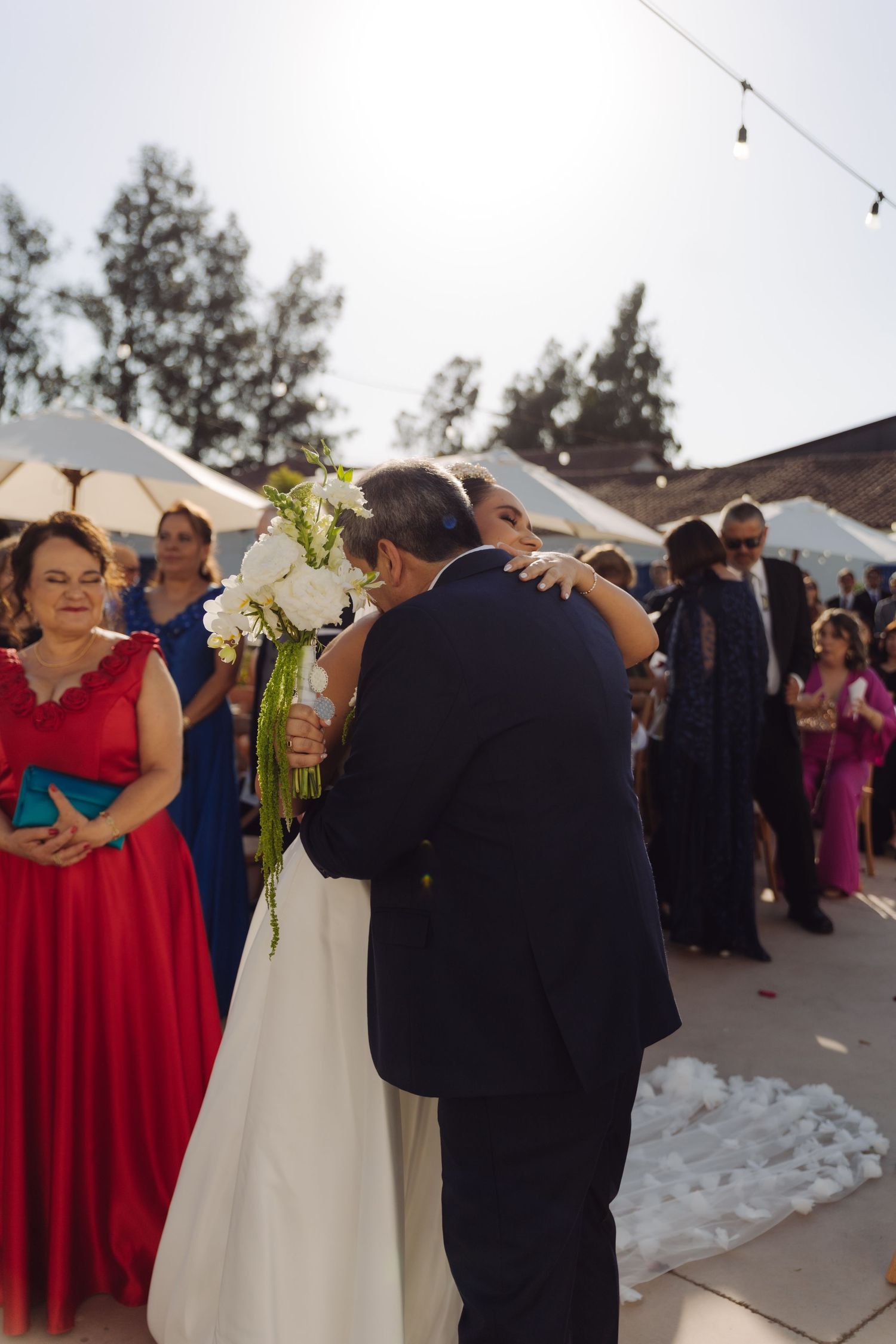 Father embraces bride in white gown during outdoor wedding ceremony processional.