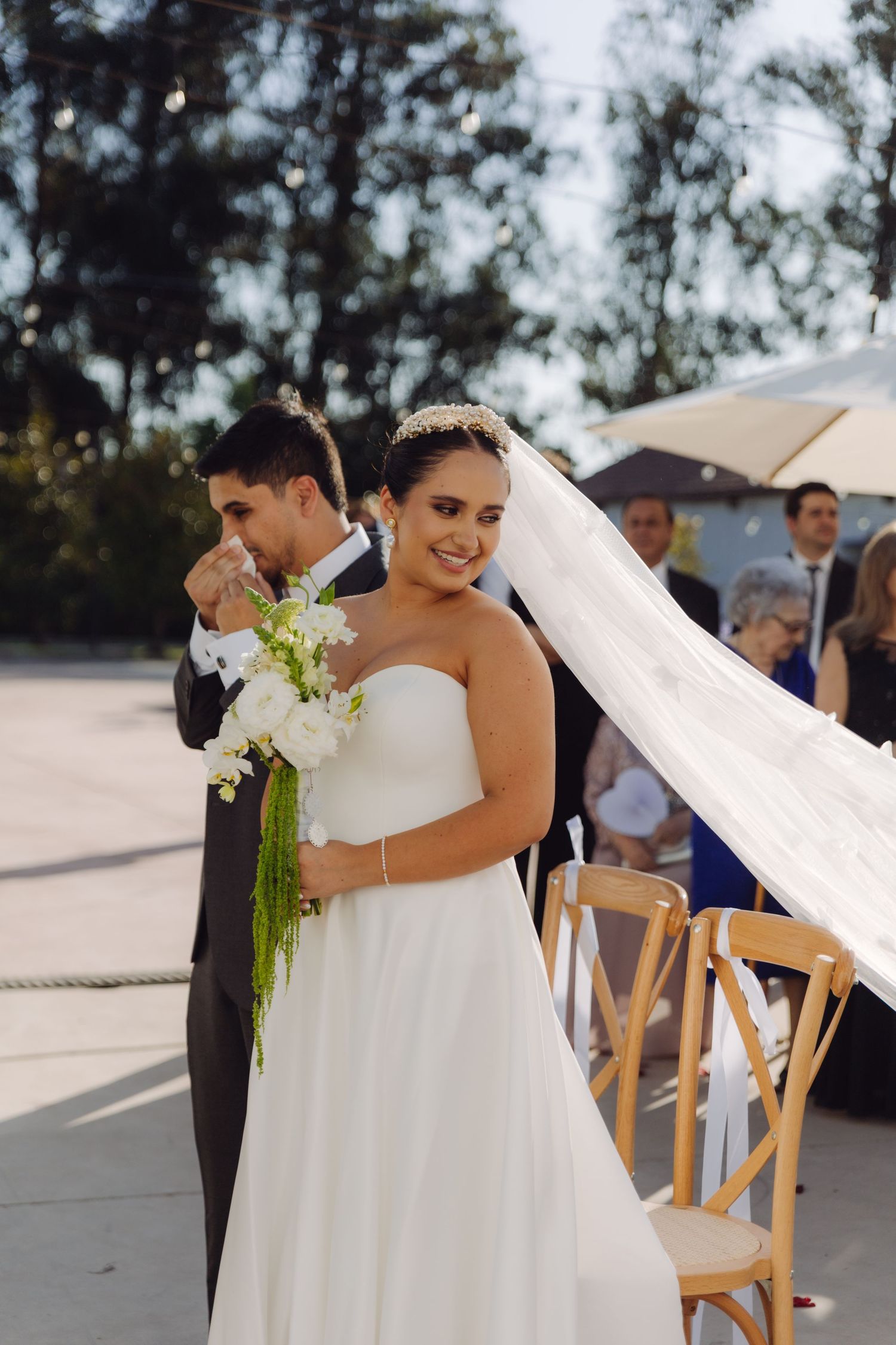 Emotional groom wipes tears as radiant bride smiles during outdoor wedding ceremony.