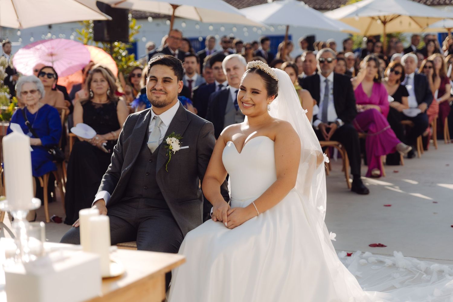 Bride and groom sit together smiling during outdoor wedding ceremony with guests in background.