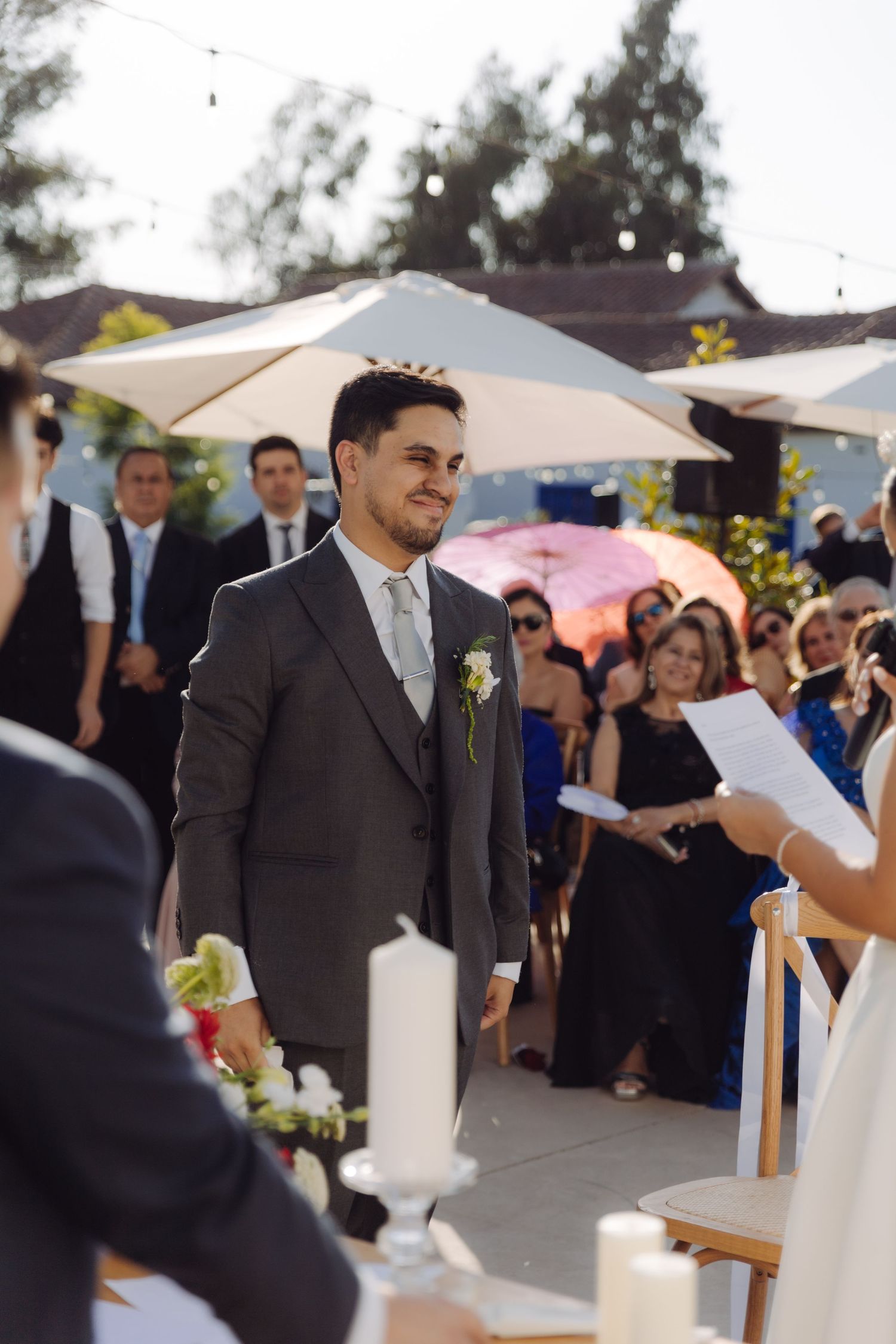 Smiling groom in gray suit stands at altar during outdoor wedding ceremony with guests seated.
