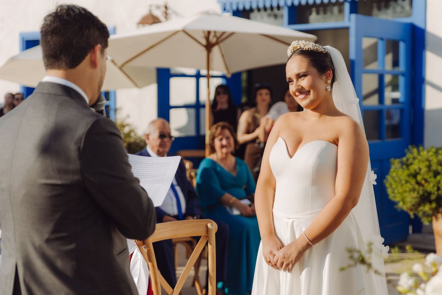 Bride in strapless white gown smiles at groom during outdoor wedding ceremony.