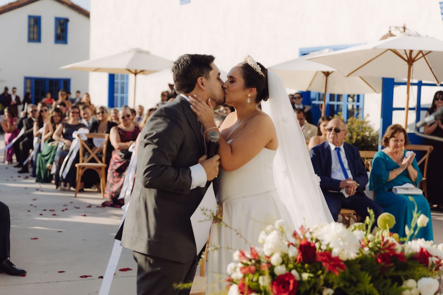 Bride and groom share a passionate first kiss at outdoor wedding ceremony with guests applauding.