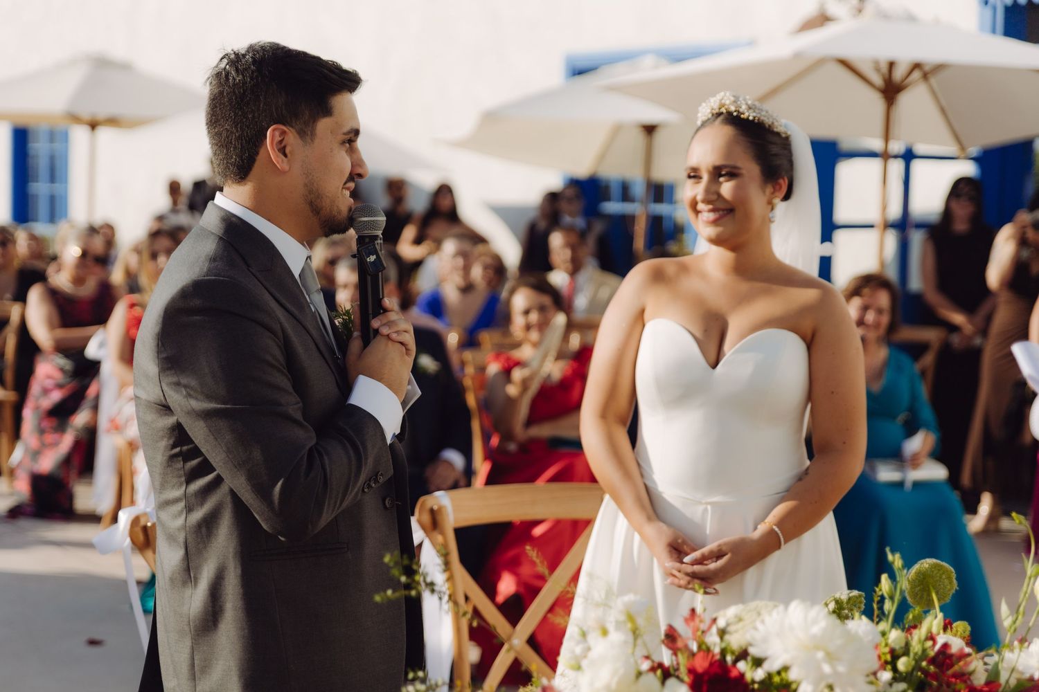 Groom speaks into microphone while bride smiles joyfully during outdoor wedding ceremony.