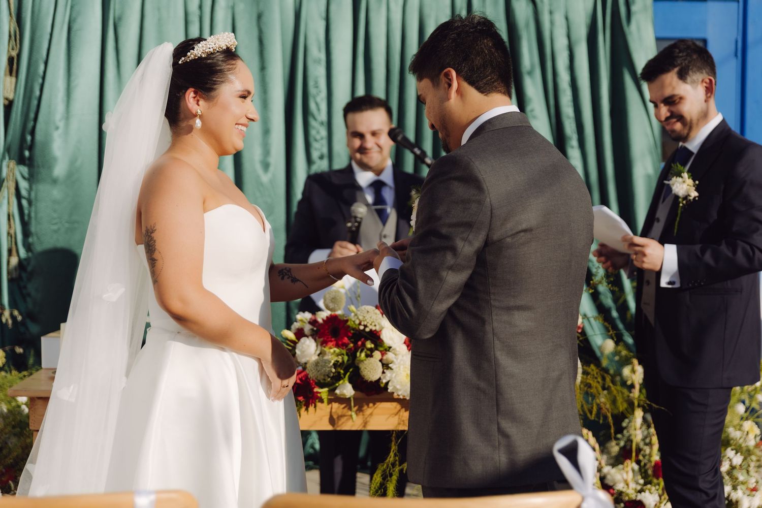 Bride and groom exchange rings during outdoor wedding ceremony with officiant and witness standing behind them.