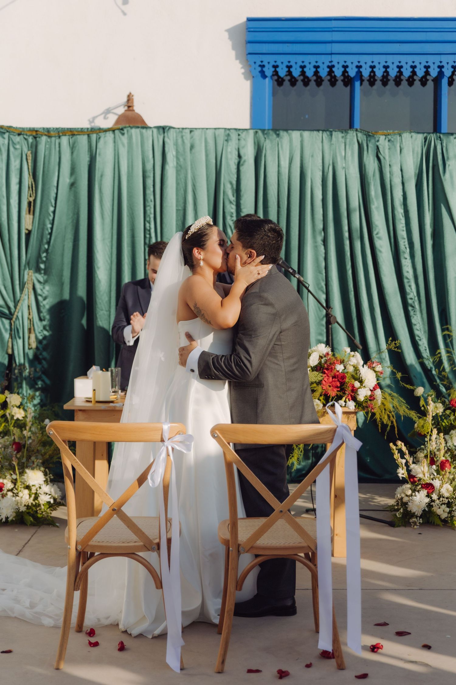 Newlyweds share first kiss at outdoor wedding ceremony altar adorned with floral arrangements and green drapes.