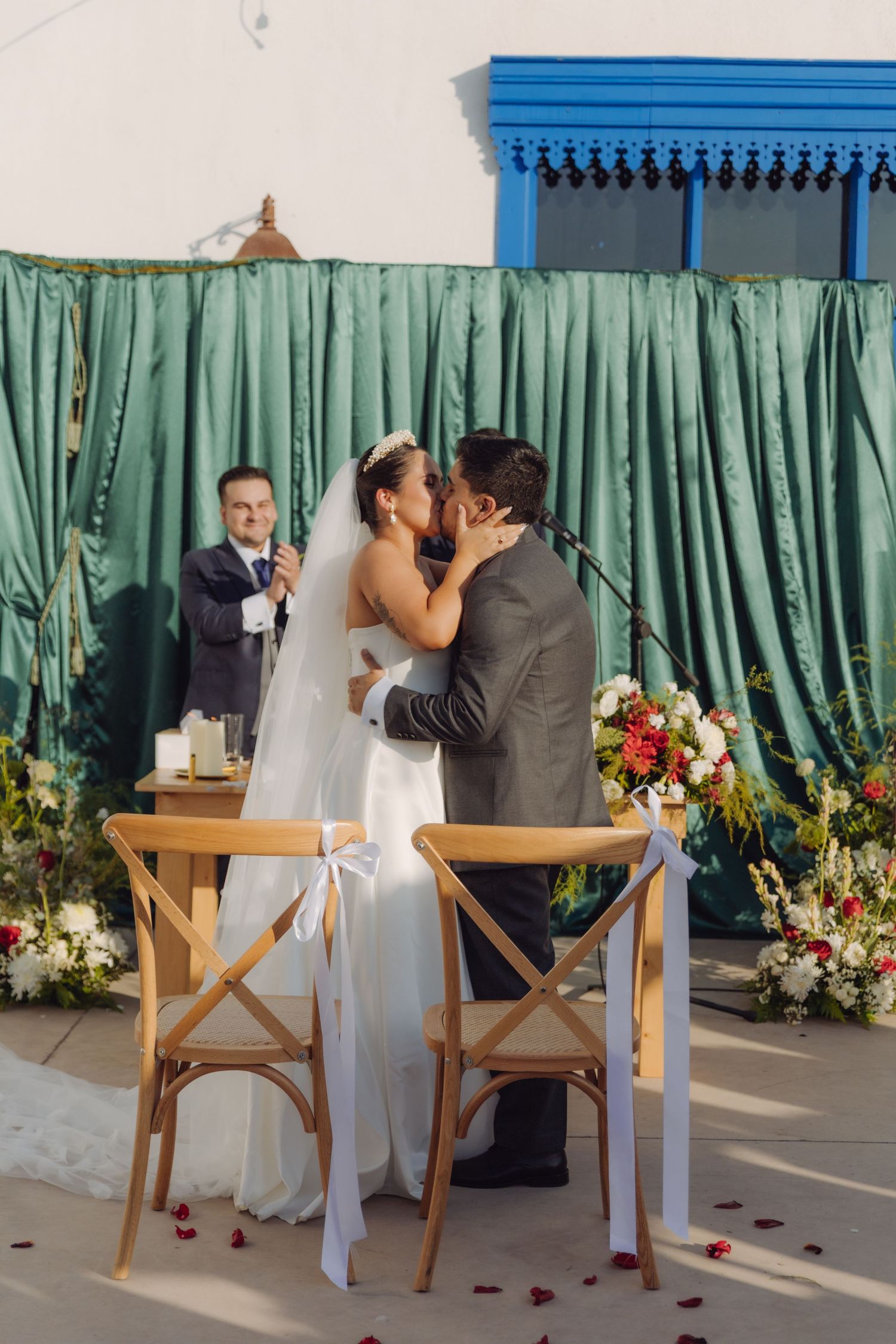 Bride and groom kiss at wedding altar while smiling officiant looks on beside green curtain backdrop.