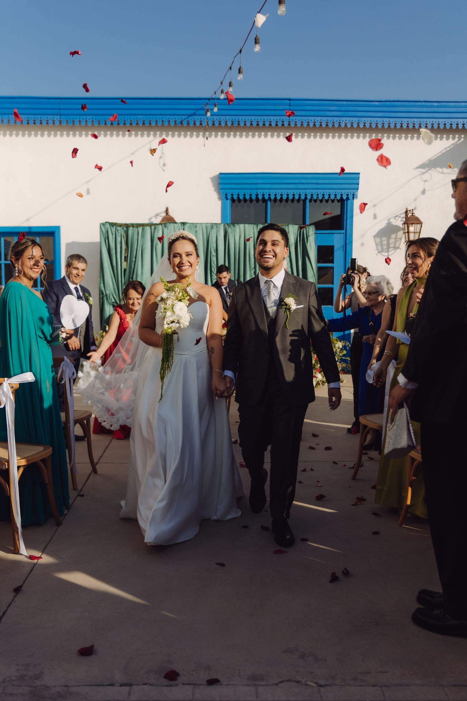 Joyful newlyweds walk down aisle as guests toss red rose petals at outdoor wedding ceremony.