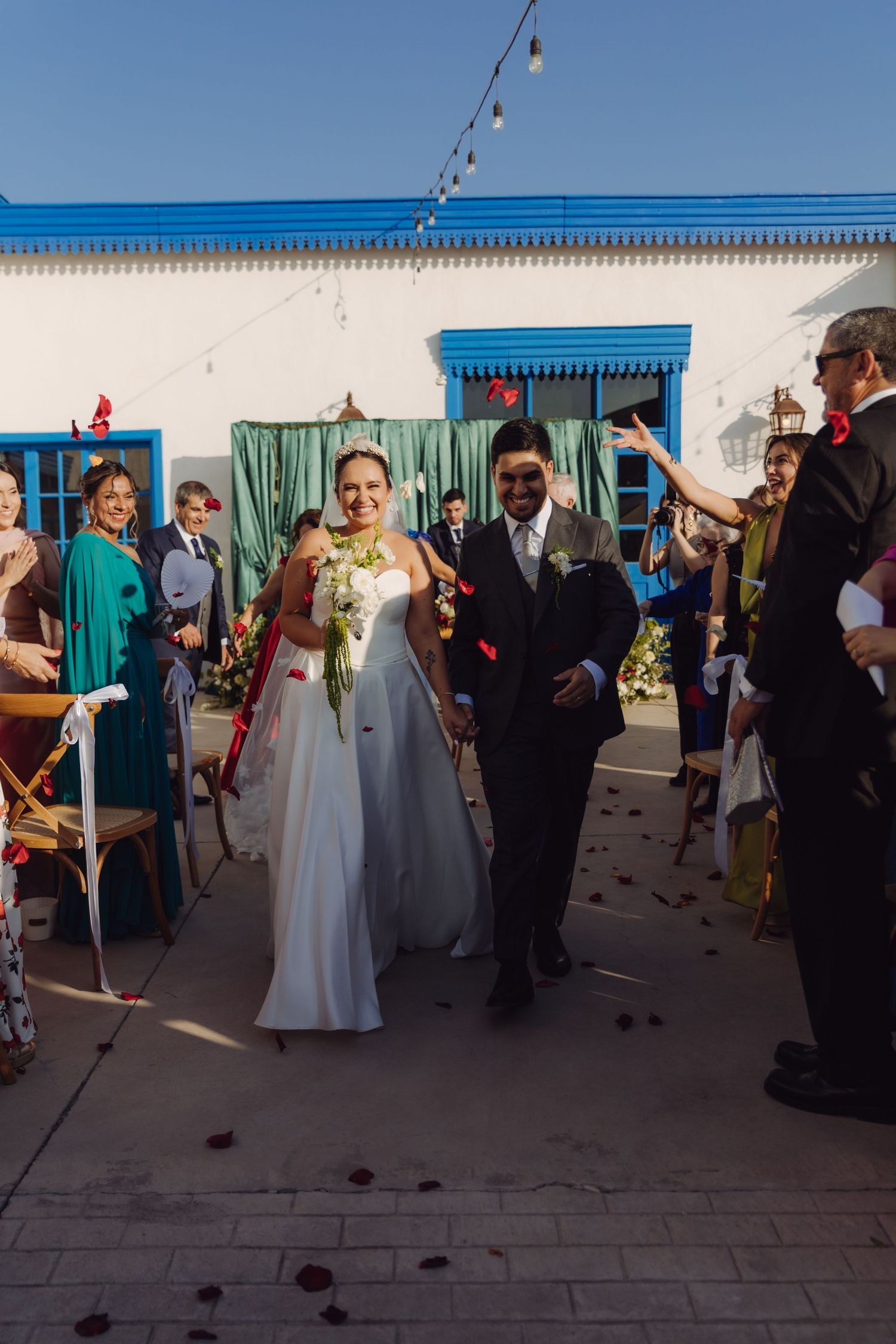 Smiling bride and groom walk through petal-throwing guests after outdoor wedding ceremony at white building venue.