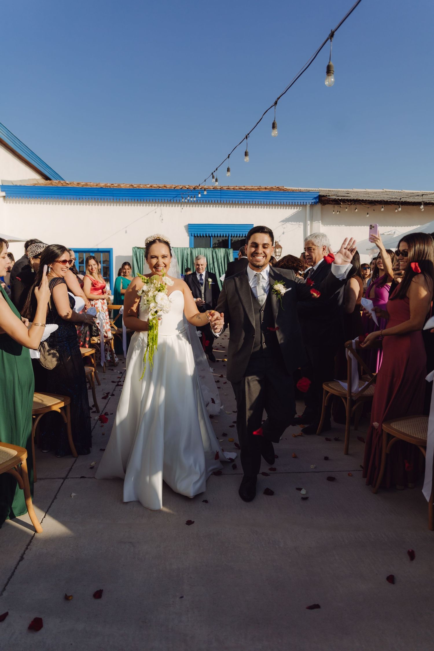 Happy bride and groom hold hands walking down aisle as guests celebrate with rose petal toss.