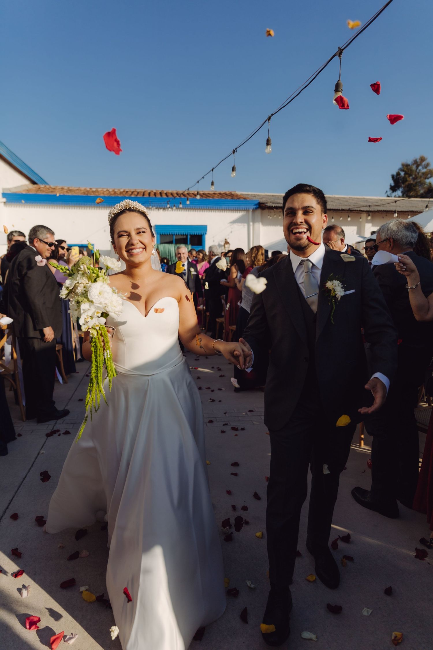 Laughing newlyweds walk through colorful confetti toss surrounded by cheering guests at outdoor wedding ceremony.