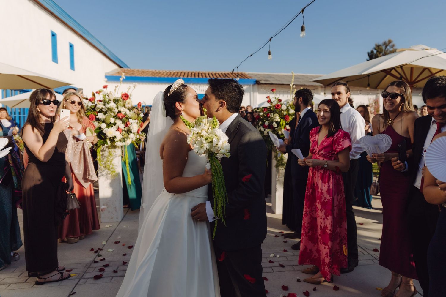 Newlyweds share romantic kiss surrounded by smiling wedding guests at outdoor ceremony with floral decorations.
