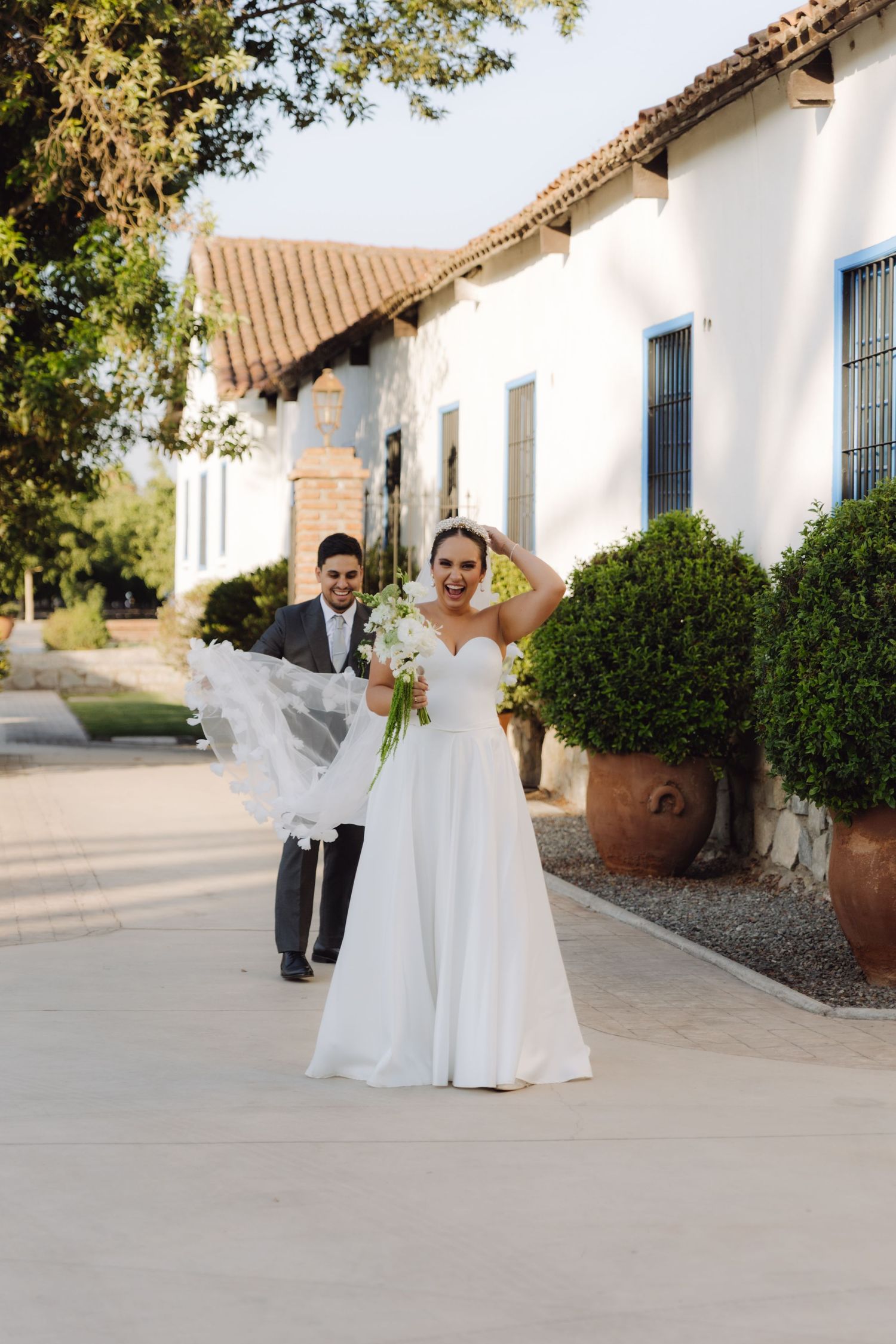 Bride in flowing white gown holds bouquet while groom adjusts her veil on sunny wedding day.