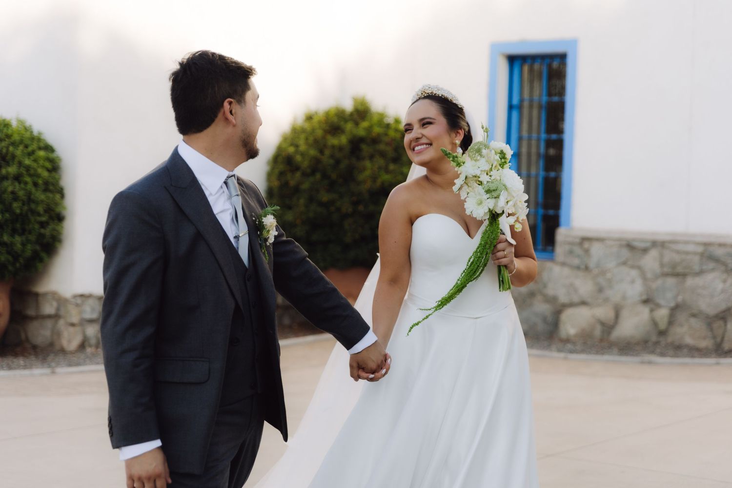 Bride and groom holding hands smiling at each other outside a white building on their wedding day.