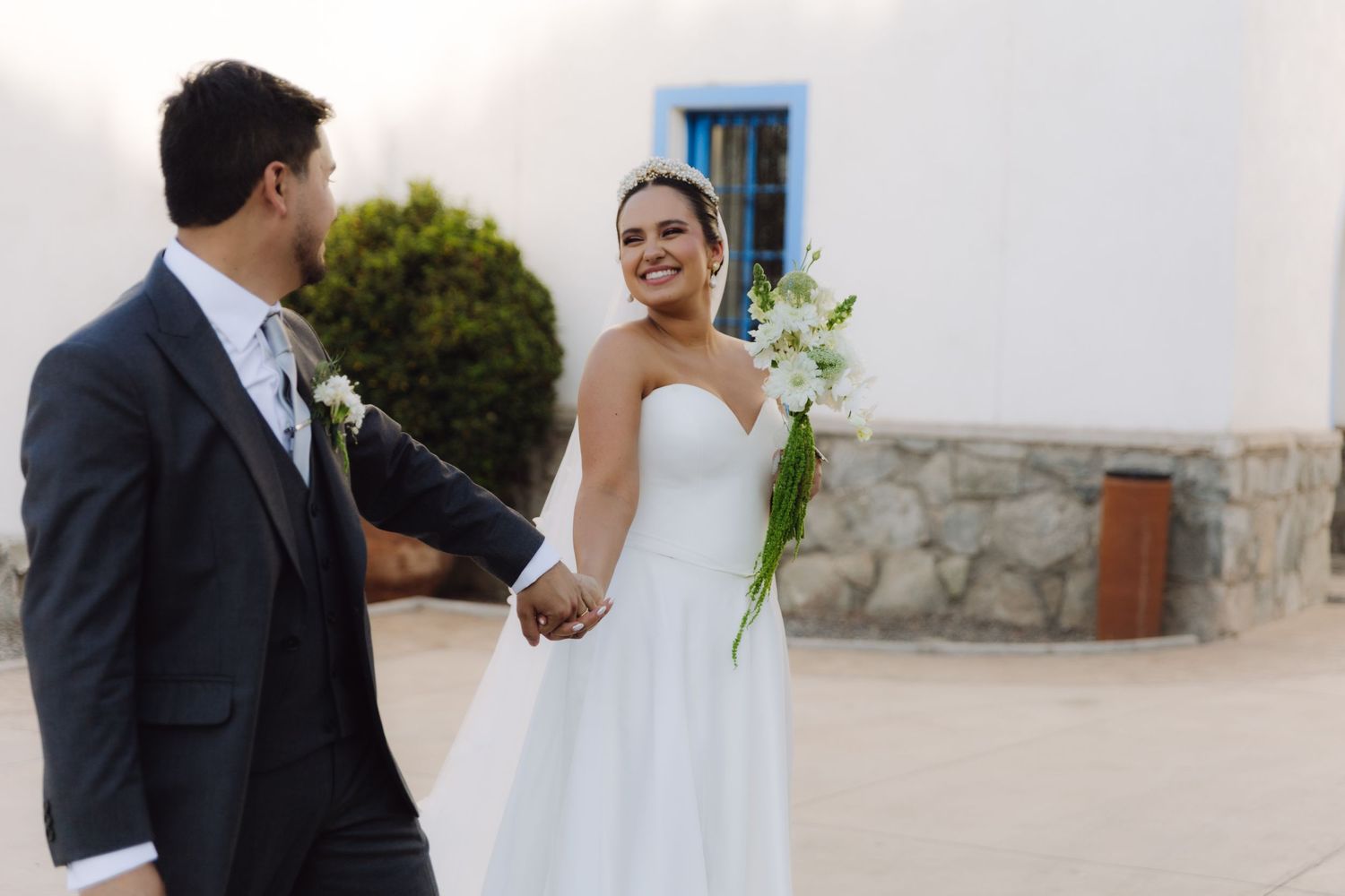 Happy bride in strapless white gown holding white bouquet walking with groom in dark suit.