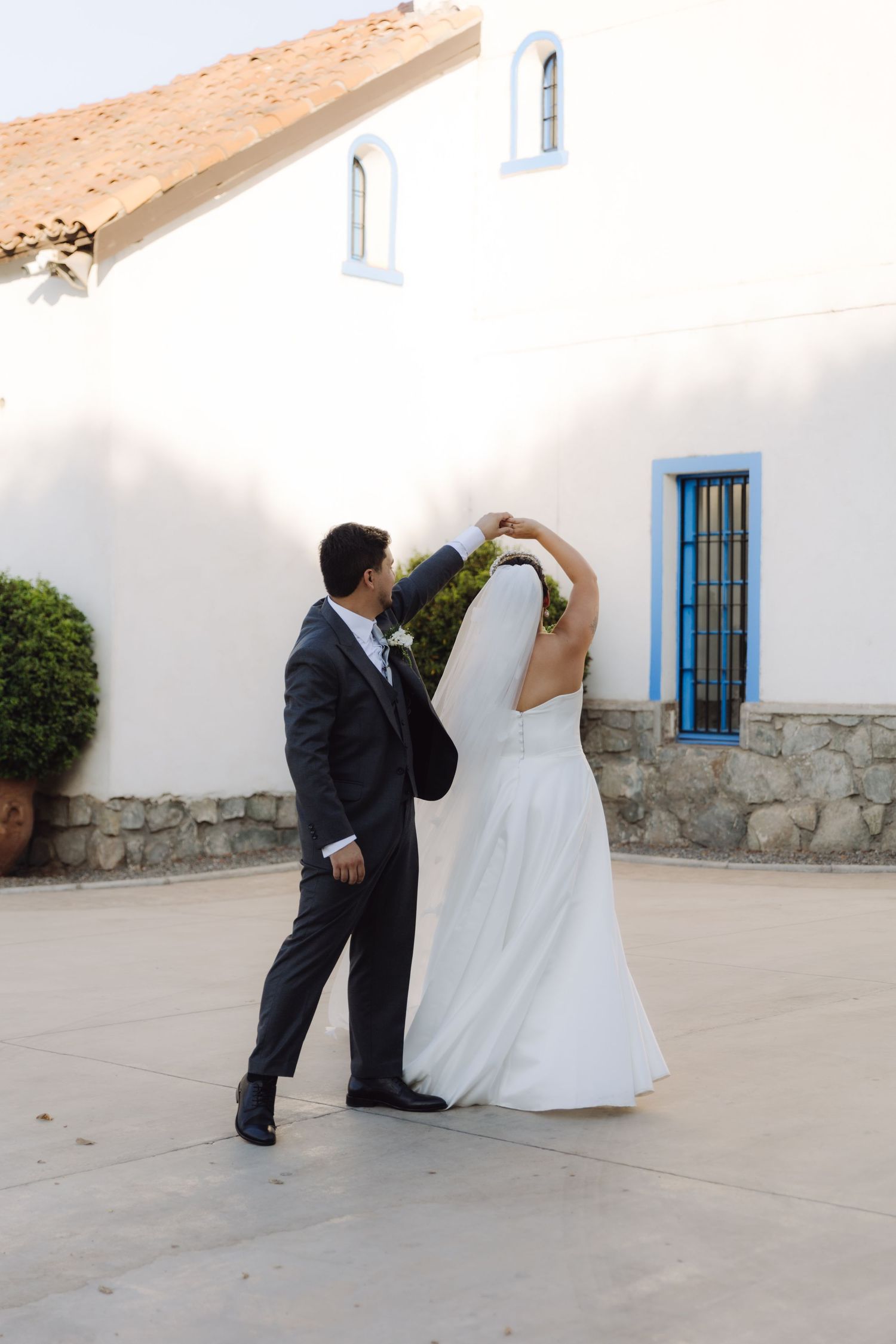 Groom spinning bride in white wedding dress outside a whitewashed building with blue windows.