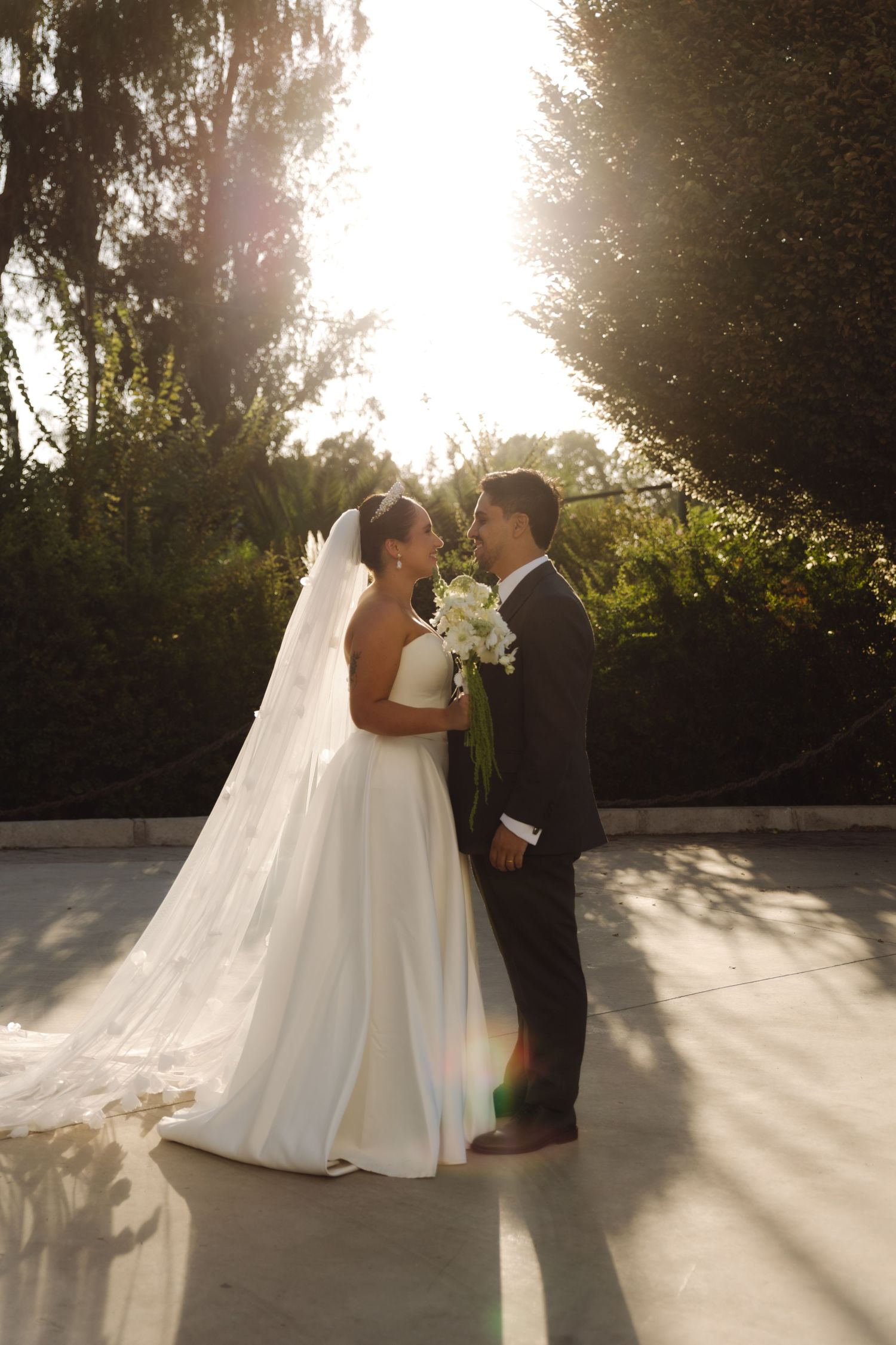Bride and groom facing each other in golden sunset light surrounded by lush green trees.