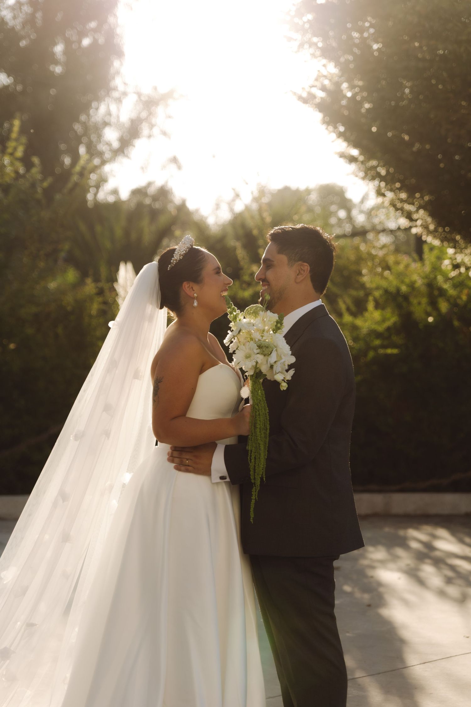 Romantic bride and groom embracing outdoors in warm sunlight with flowing veil and white bouquet.