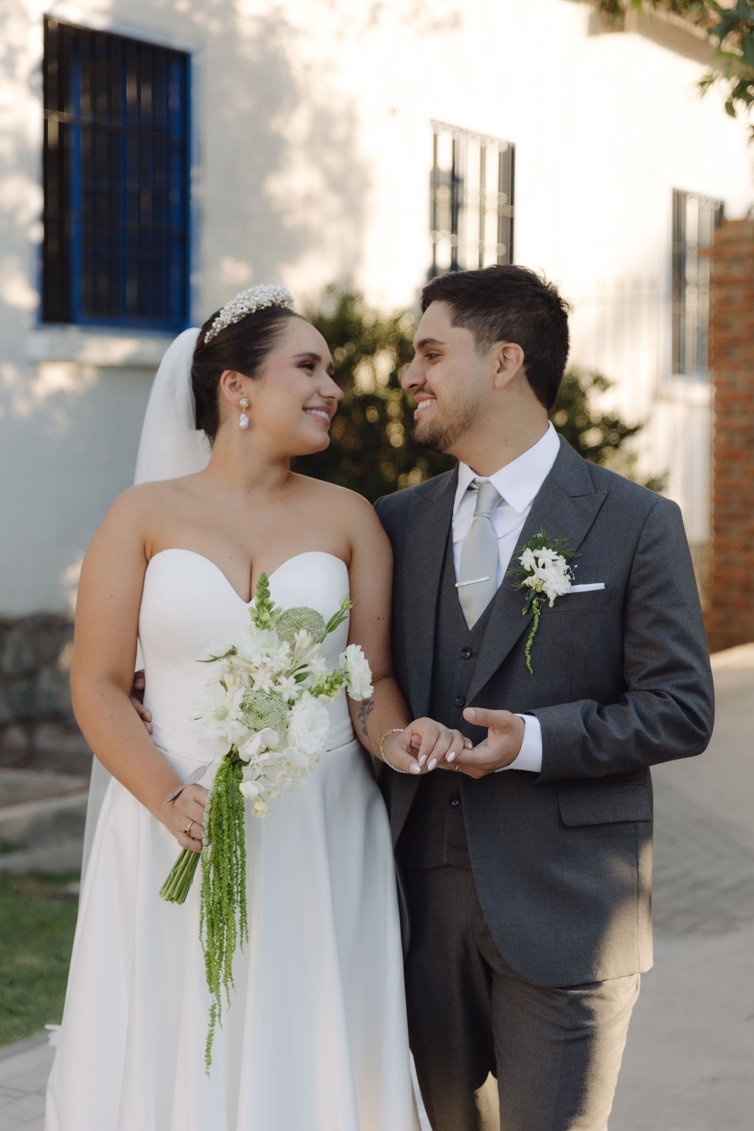 Smiling bride and groom walking together holding hands near a white building.