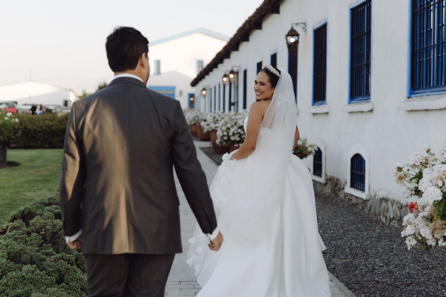 Bride and groom walking hand in hand along a path beside a white building.