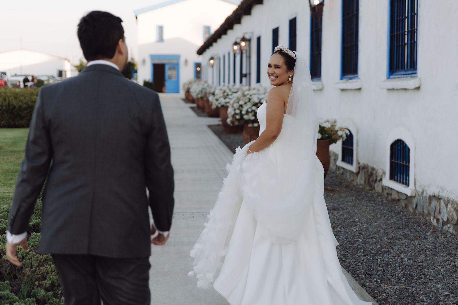 Bride smiling back at groom while walking along a white building pathway.