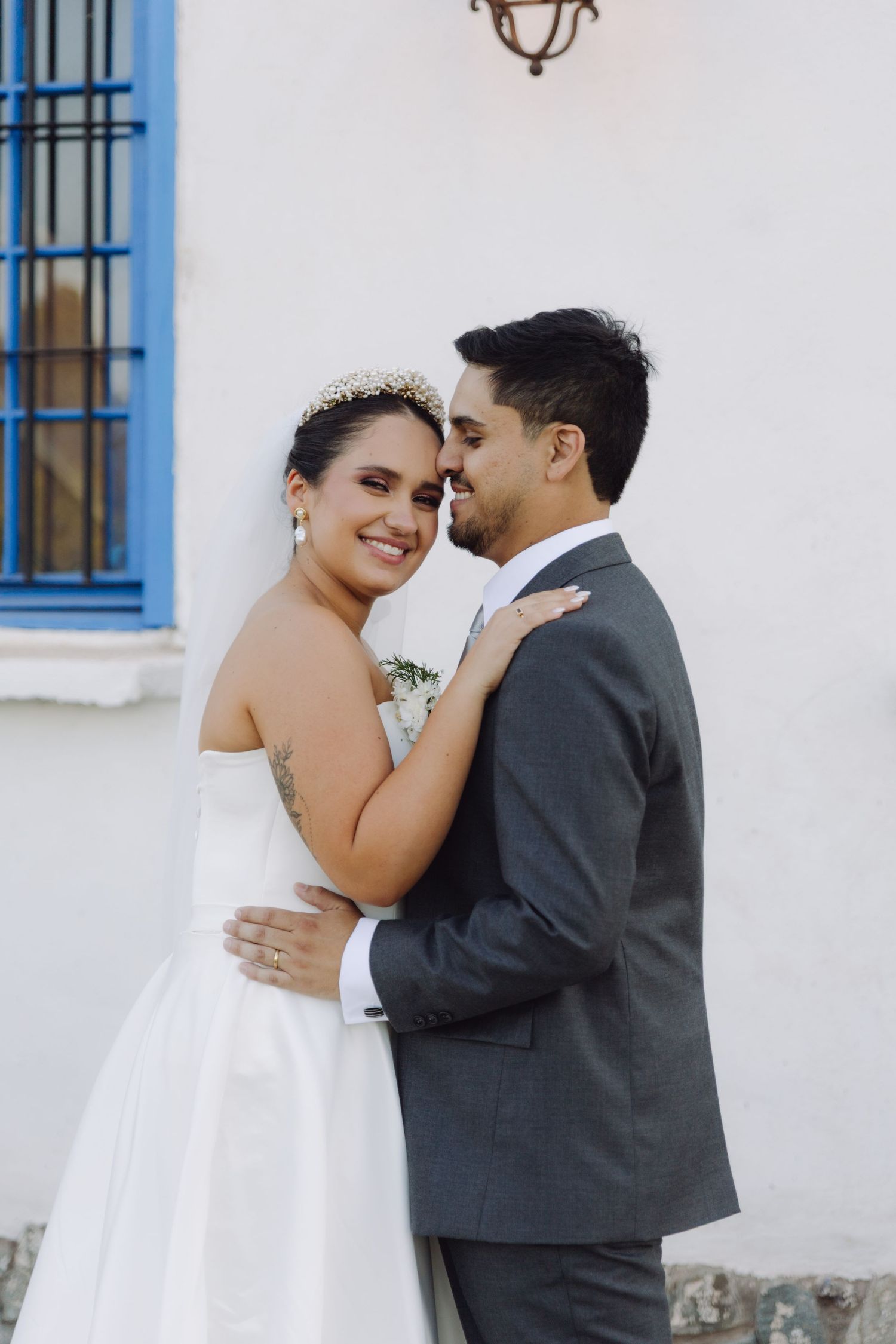 Bride in strapless white gown and groom in gray suit embrace and smile against a white wall with blue window.