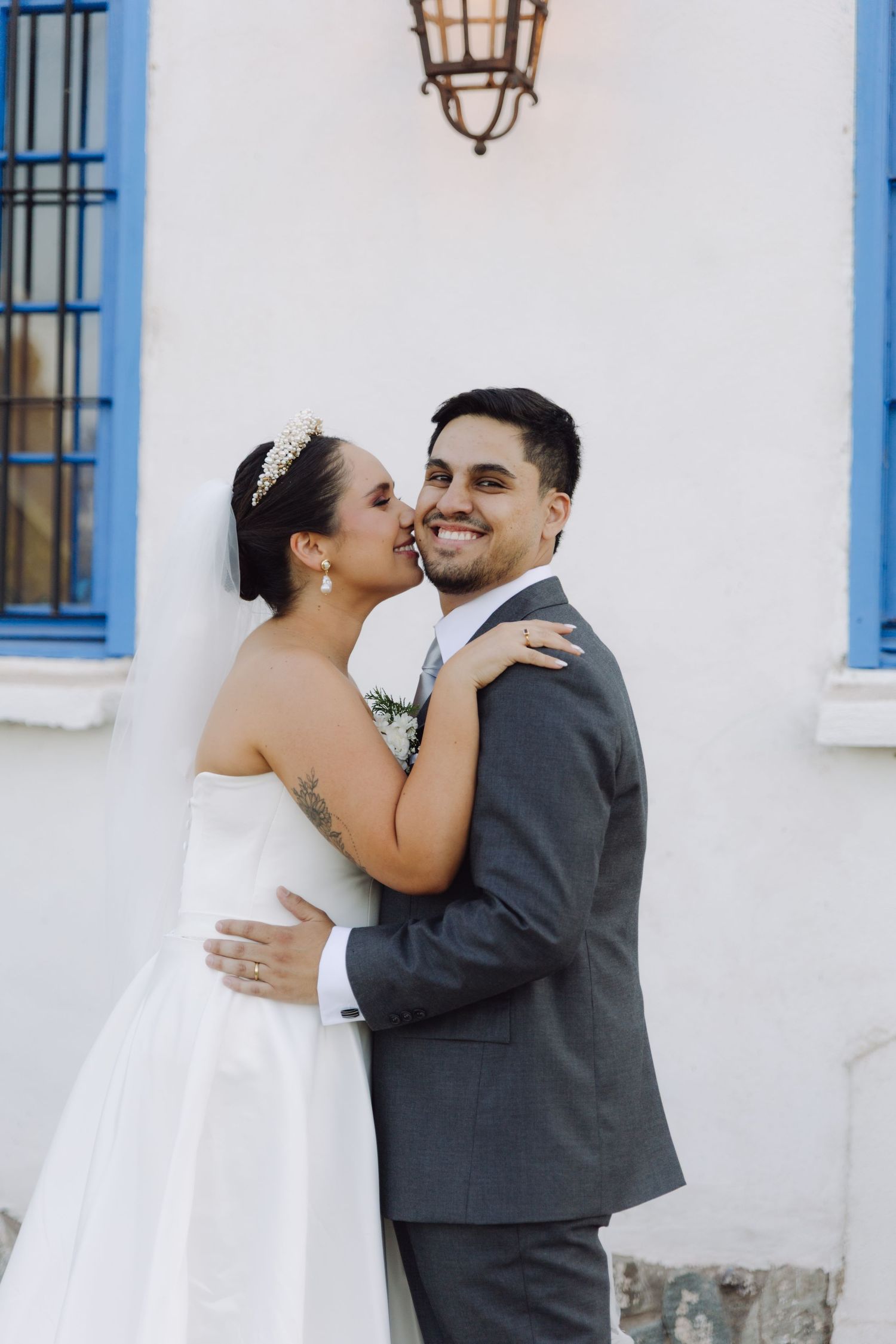 Bride whispers to smiling groom in gray suit, embracing outdoors near a white building with blue window.