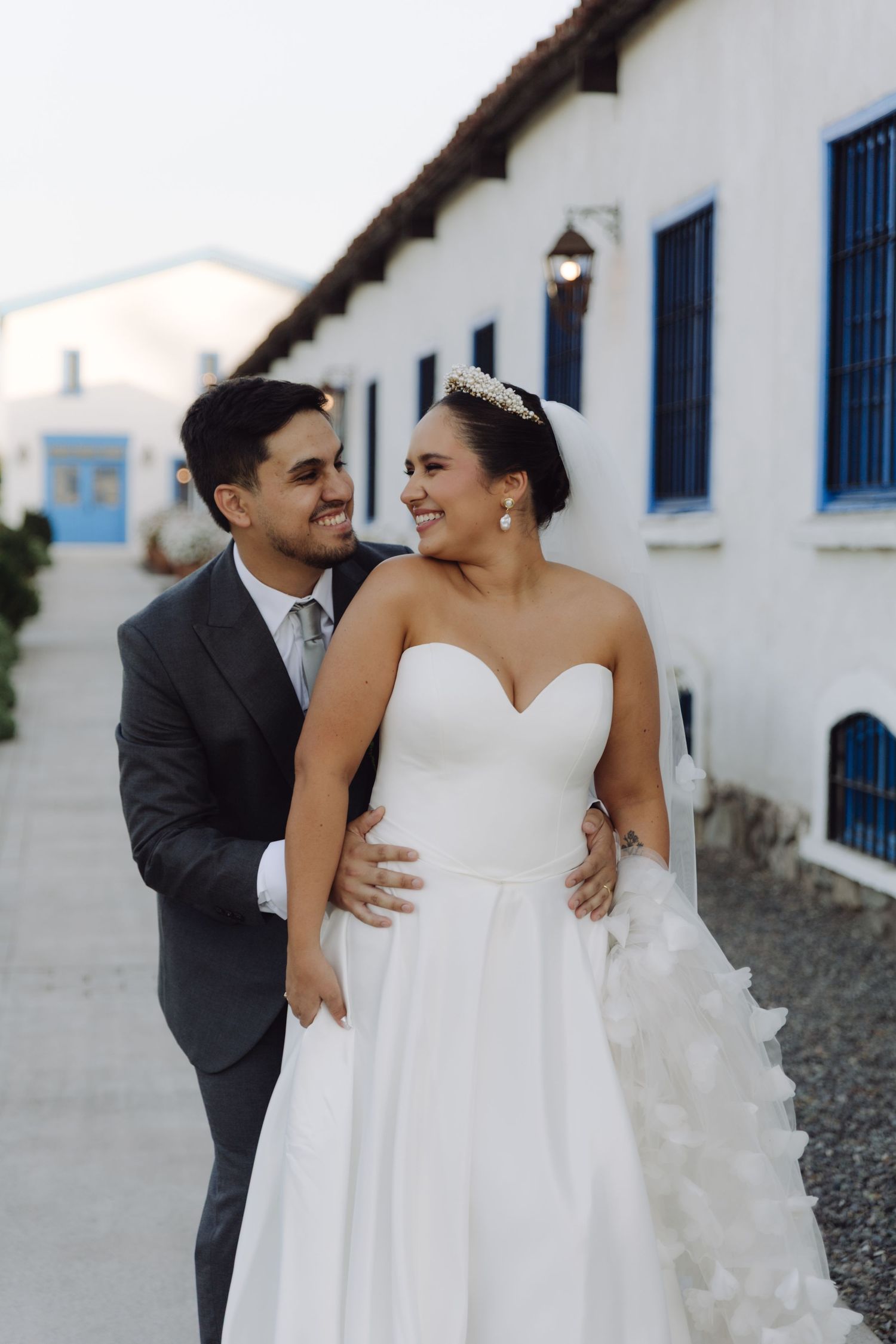 Bride in sweetheart ballgown and groom in gray suit walk together laughing outside a white building with blue shutters.