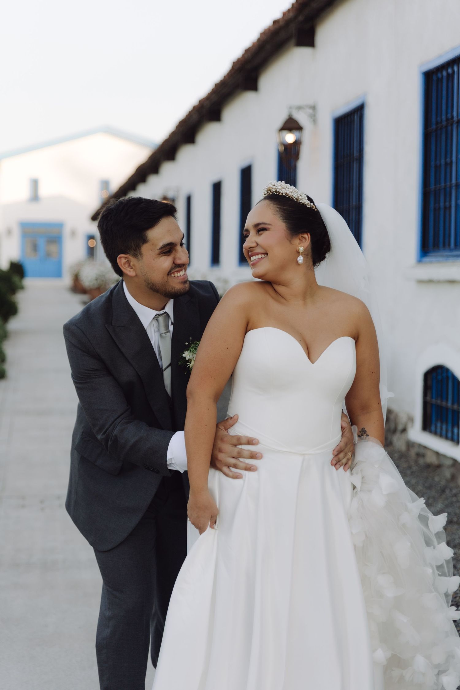 Happy bride in white ballgown and groom in gray suit share a joyful moment outside a white building with blue shutters.