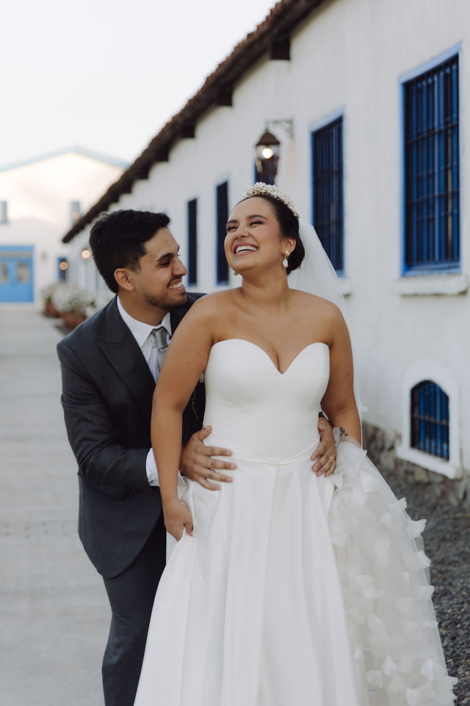 Bride in strapless white ballgown laughs as groom in gray suit holds her waist outside a white building at dusk.