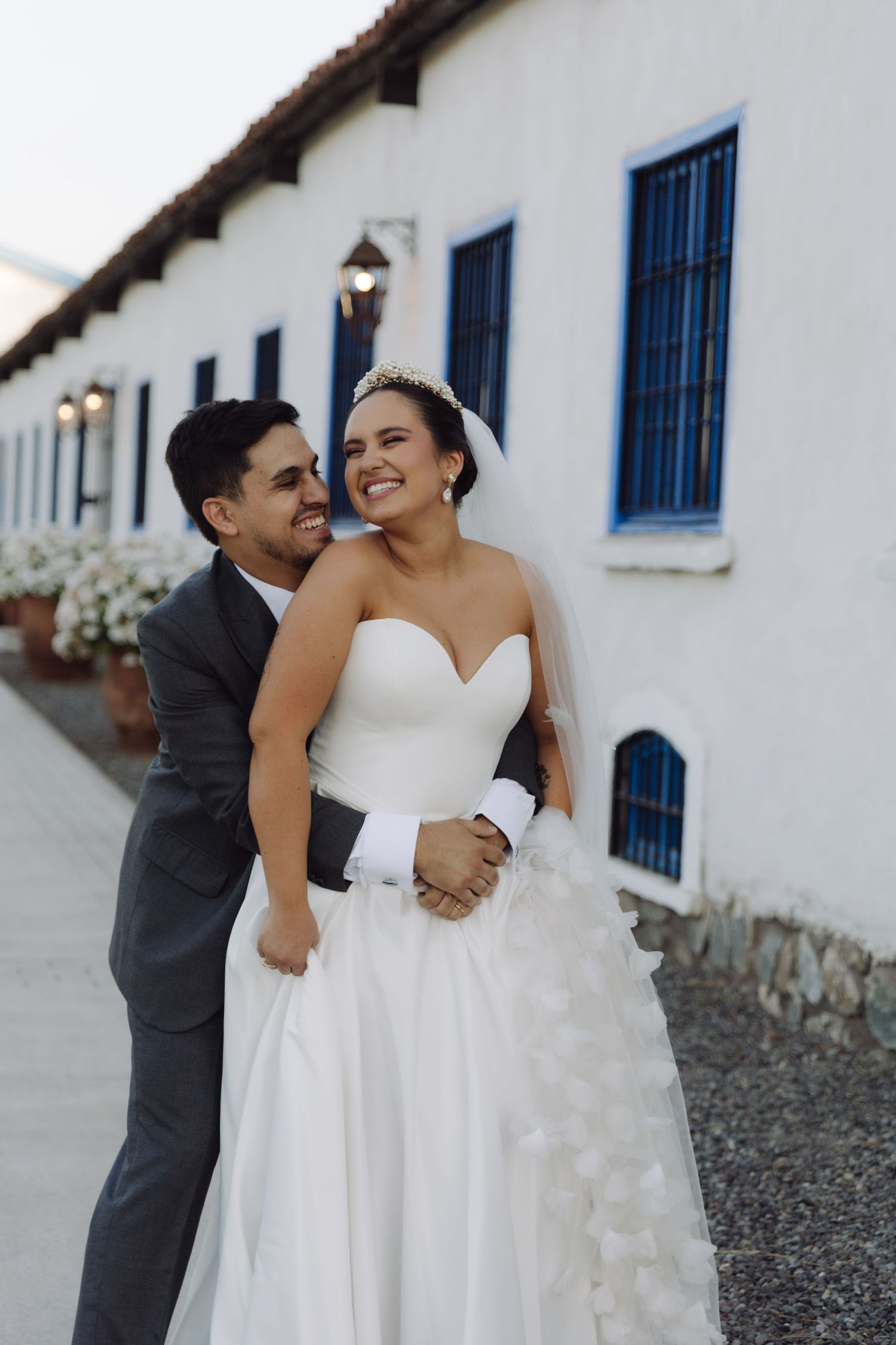 Groom in gray suit embraces bride in white ballgown from behind as they smile outside a white building at dusk.