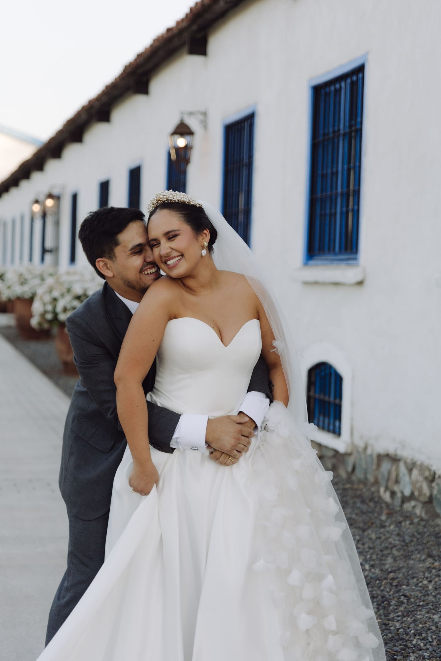 Groom nuzzles smiling bride in strapless white ballgown from behind outside a white building with blue shutters.