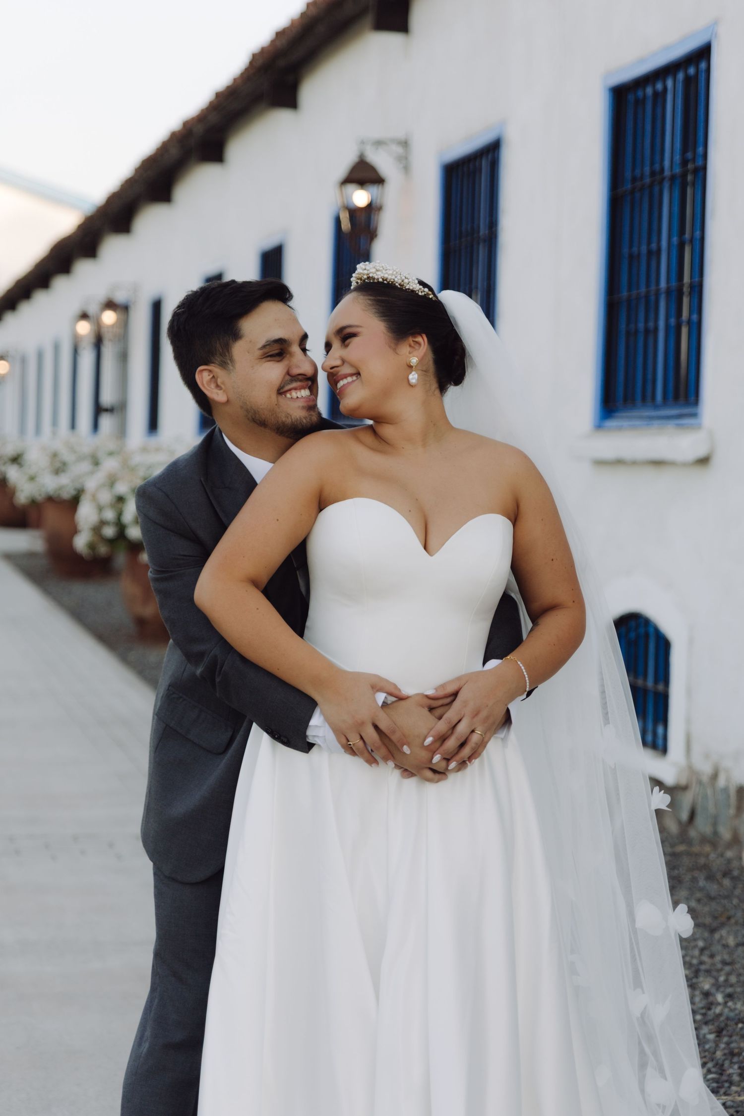 Bride and groom gaze at each other tenderly, embracing in front of a white building with blue windows at dusk.