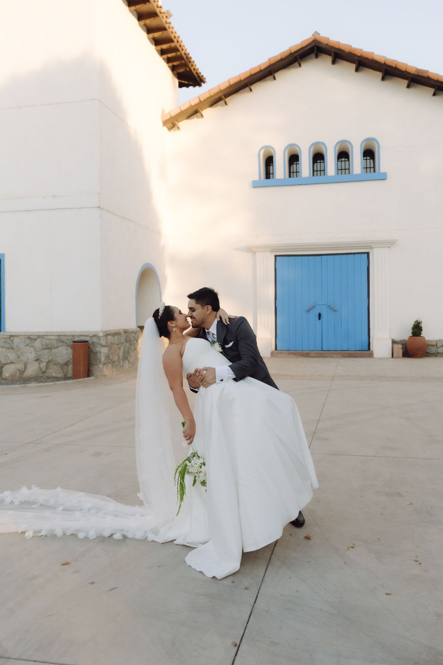 Groom dips bride in white gown and veil for a kiss in front of a white chapel with blue doors.