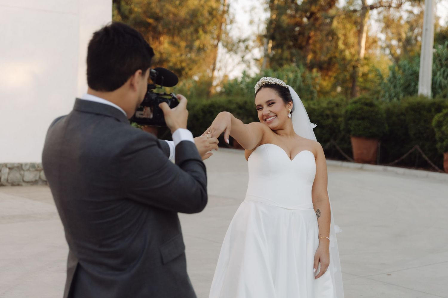 Groom photographs smiling bride in strapless wedding dress outdoors.