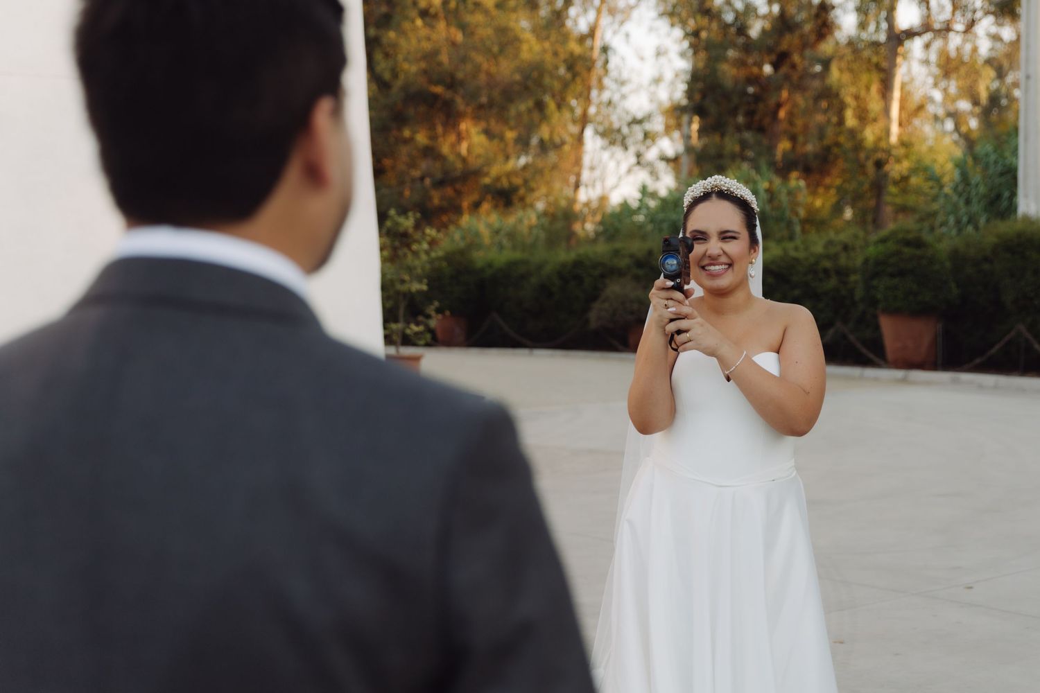 Smiling bride photographs groom in gray suit during outdoor wedding shoot.