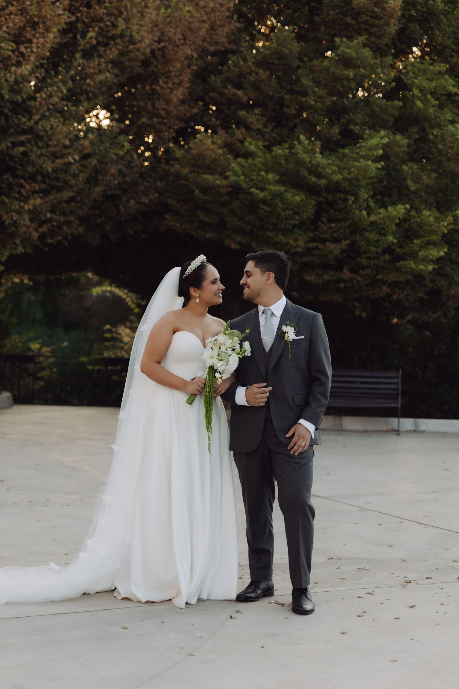 Bride and groom share a loving glance holding a white bouquet outdoors.