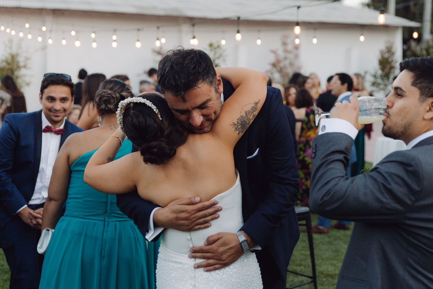 Groom embraces bride warmly surrounded by wedding guests at reception.