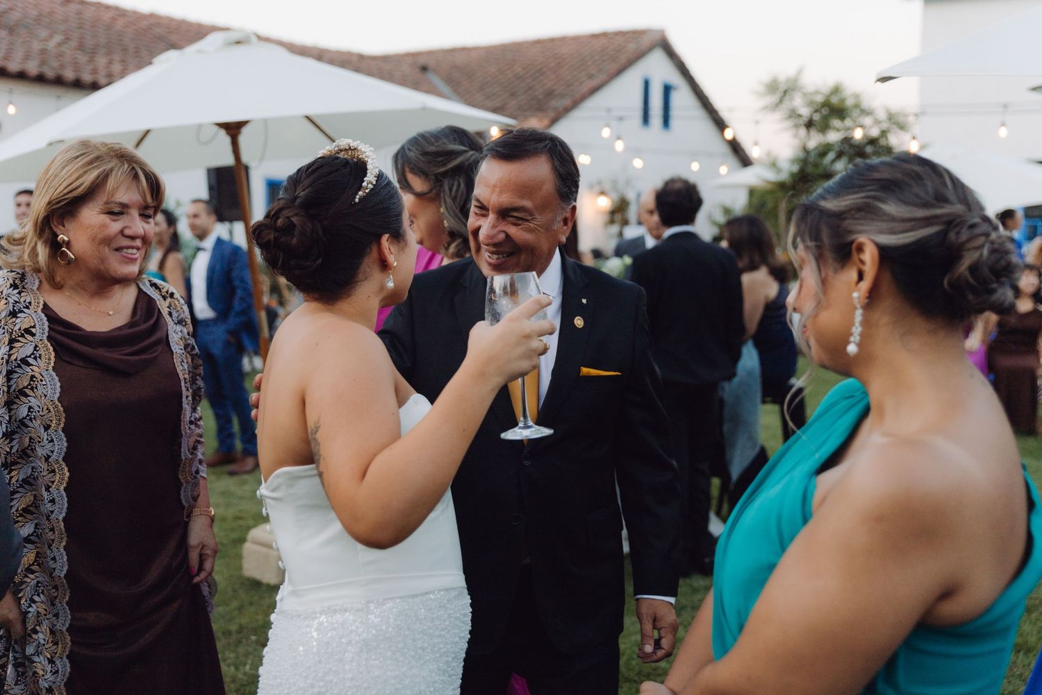 Bride in white gown toasts champagne with a smiling man in a suit at an outdoor wedding reception.