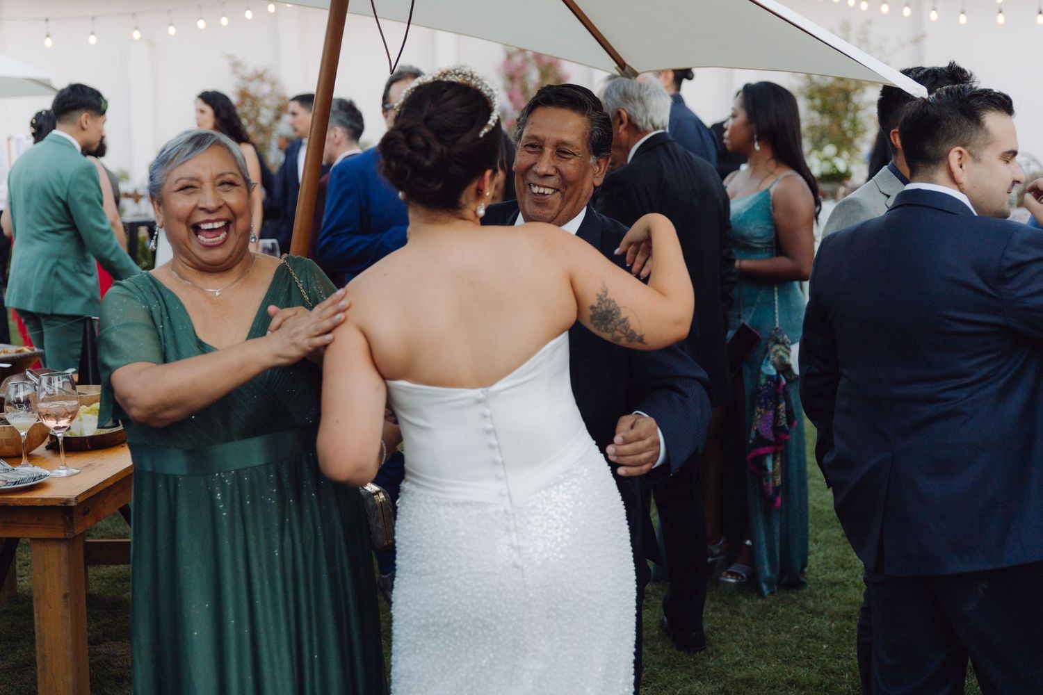 Bride in white gown laughs joyfully while hugging a man at an outdoor wedding reception.