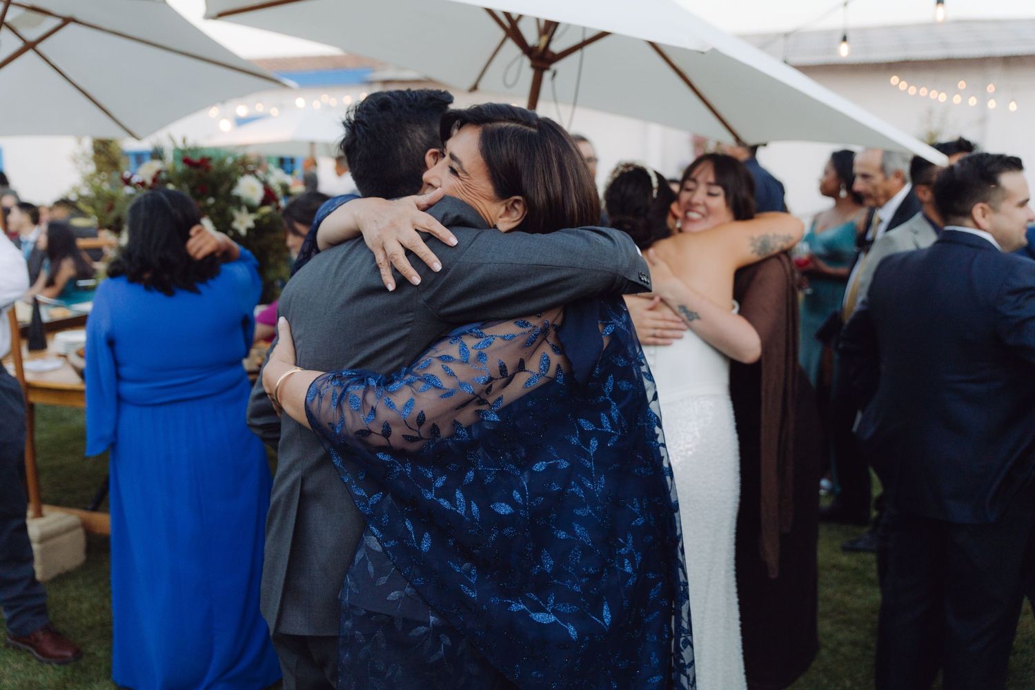 Bride and groom share a group hug with a guest in blue at an outdoor wedding reception.