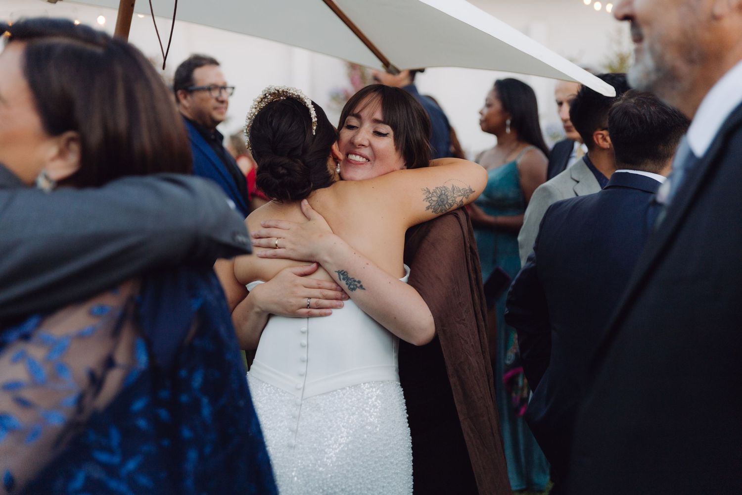 Bride in a white fitted gown smiles while embracing a guest at an outdoor wedding reception.