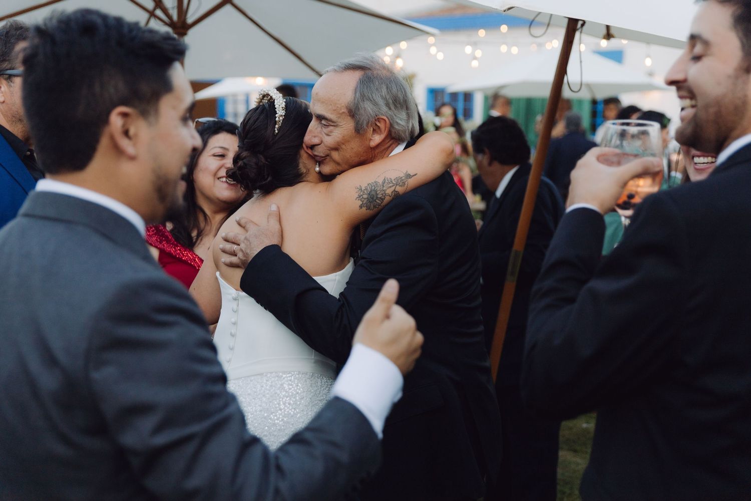 Bride in white gown receives a warm hug from an older male guest at an outdoor wedding.