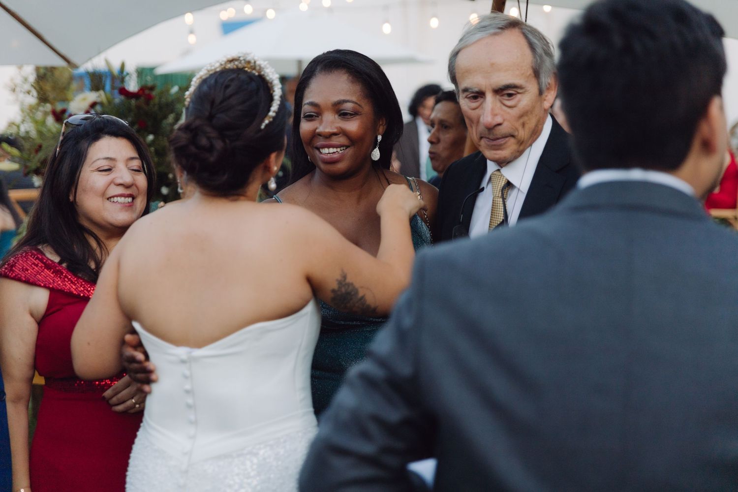 Bride in white lace gown greets smiling guests including a woman in teal at a wedding reception.