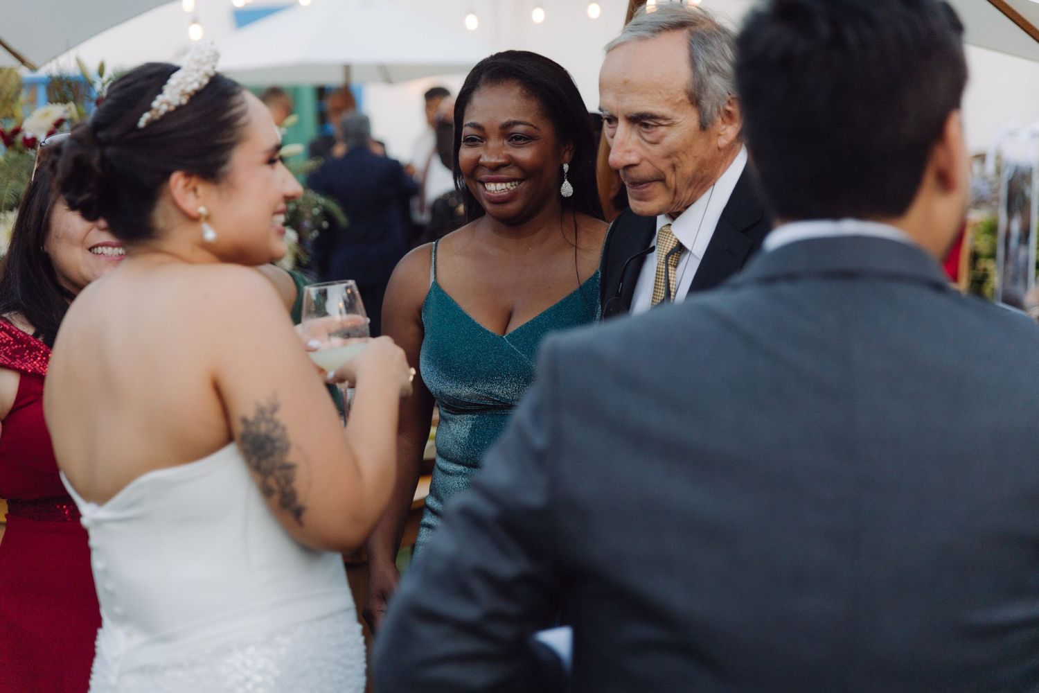 Bride in white gown chats and laughs with guests including a woman in teal at a wedding reception.